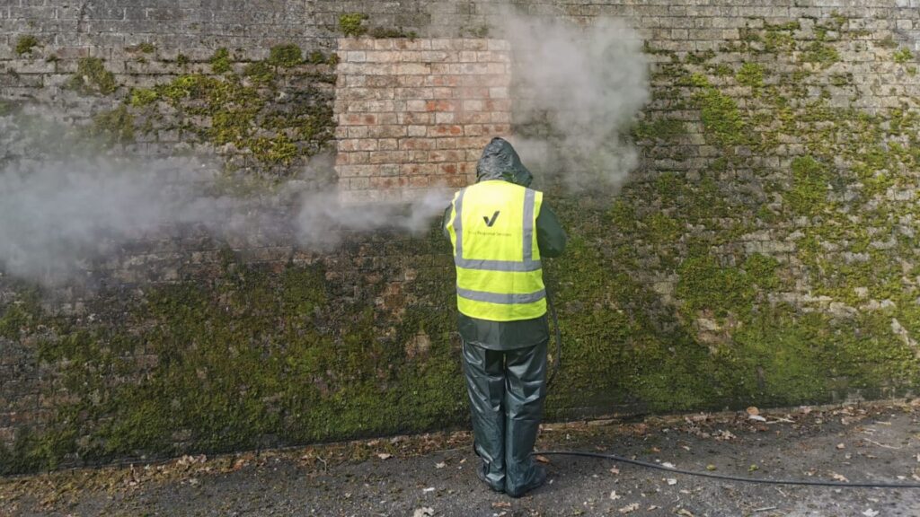 A man in a high vis jacket cleaning a dirty brick wall.