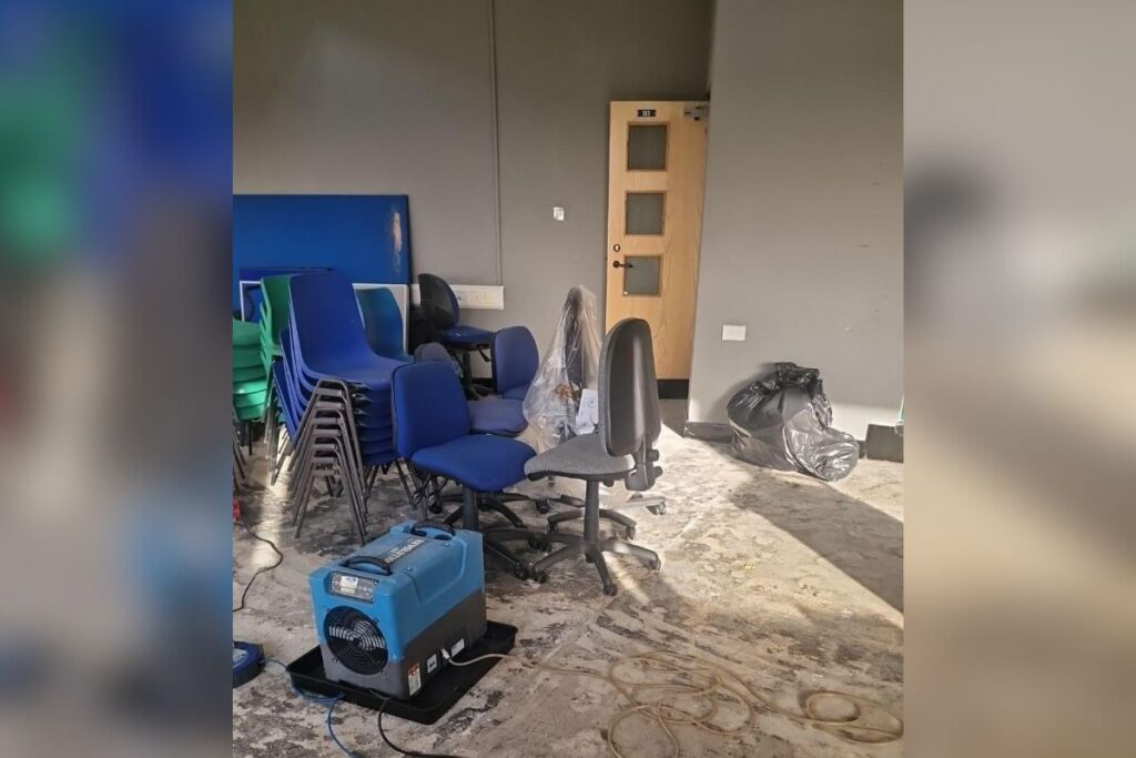 Water-damaged classroom with flooring exposed, chairs stacked, and drying equipment in operation.